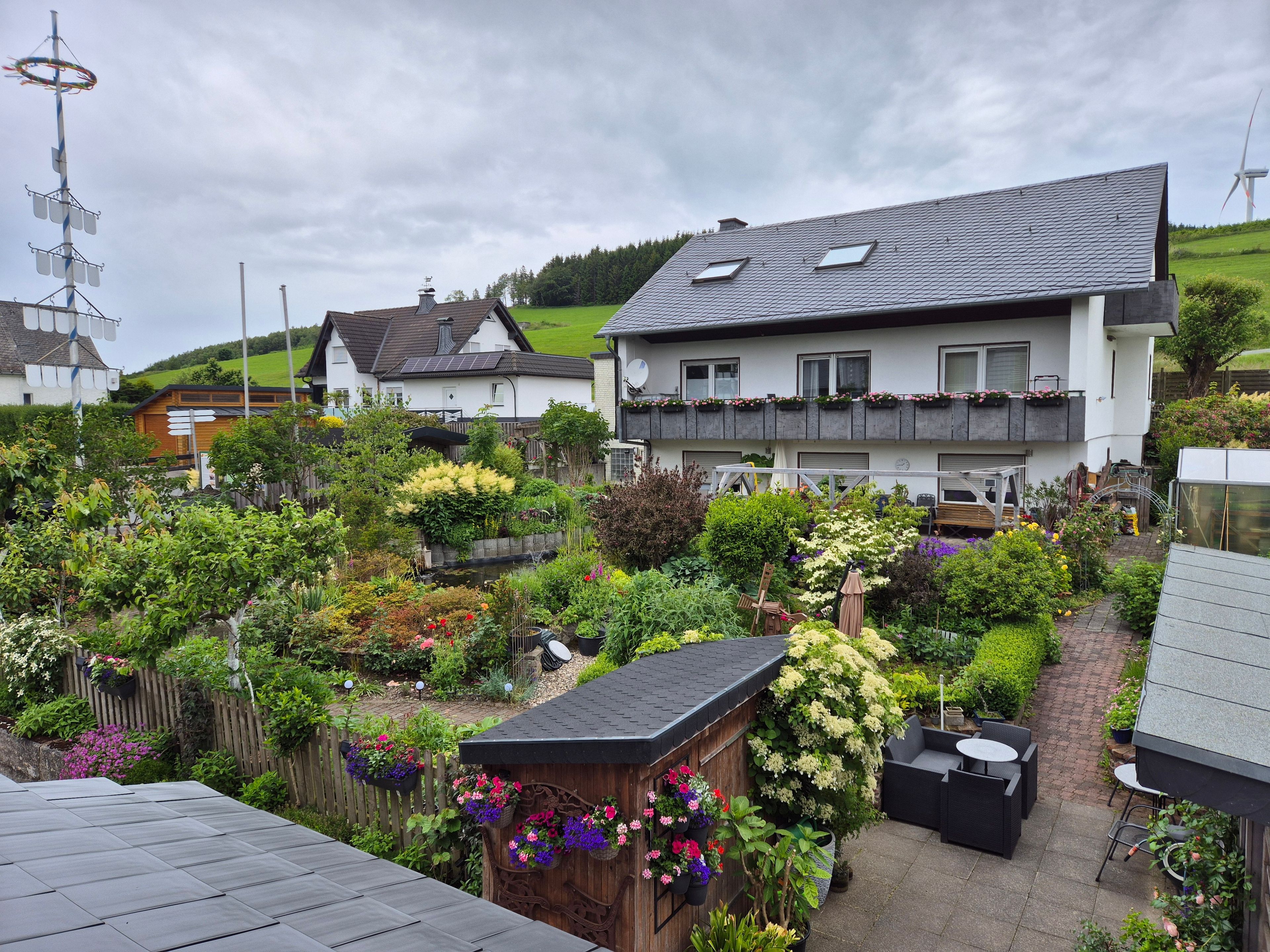 Ein Wohngebiet mit einem großen Haus, das ein geneigtes Dach, einen Garten mit verschiedenen Pflanzen und Blumen sowie eine kleine Holzhütte mit hängenden Blumenkörben zeigt.