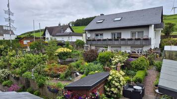 Ein Wohngebiet mit einem großen Haus, das ein geneigtes Dach, einen Garten mit verschiedenen Pflanzen und Blumen sowie eine kleine Holzhütte mit hängenden Blumenkörben zeigt.