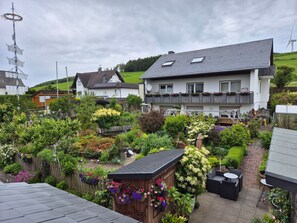 Ein Wohngebiet mit einem großen Haus, das ein geneigtes Dach, einen Garten mit verschiedenen Pflanzen und Blumen sowie eine kleine Holzhütte mit hängenden Blumenkörben zeigt.