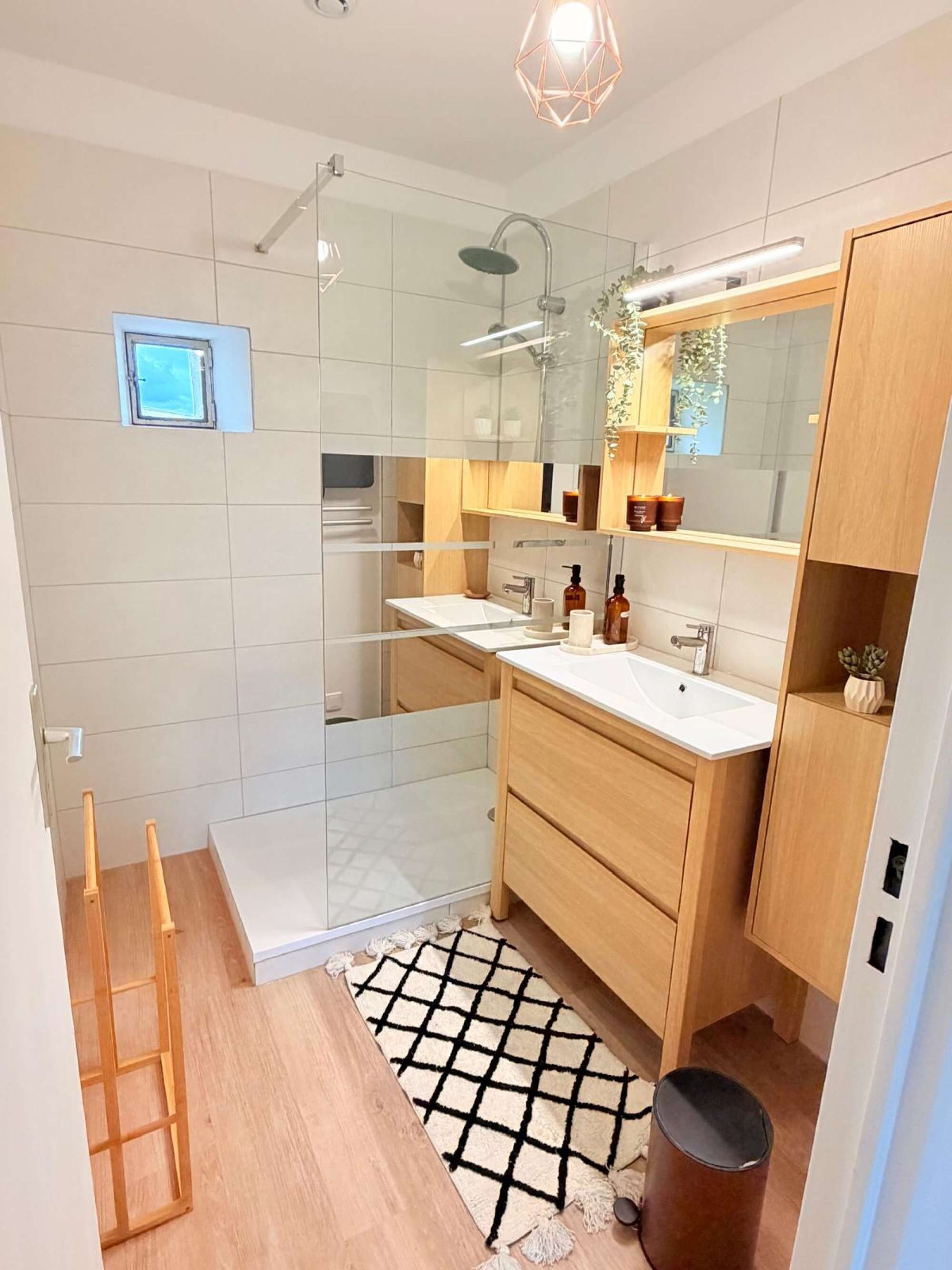 Een moderne badkamer met een douchecabine van glas, een witte wastafel, houten kastjes en een klein raam.