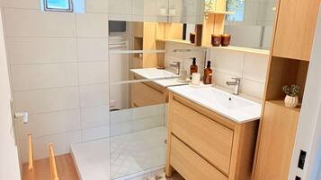 Een moderne badkamer met een douchecabine van glas, een witte wastafel, houten kastjes en een klein raam.