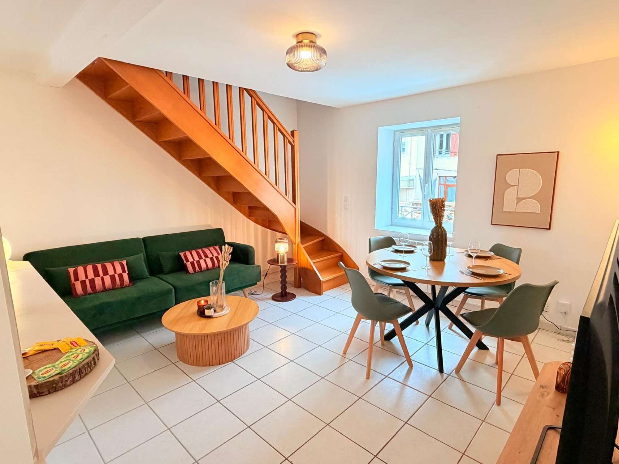 A modern living space with a green sofa, a wooden staircase, a dining area with a round table and chairs, and a large window providing natural light.