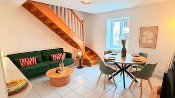 A modern living space with a green sofa, a wooden staircase, a dining area with a round table and chairs, and a large window providing natural light.