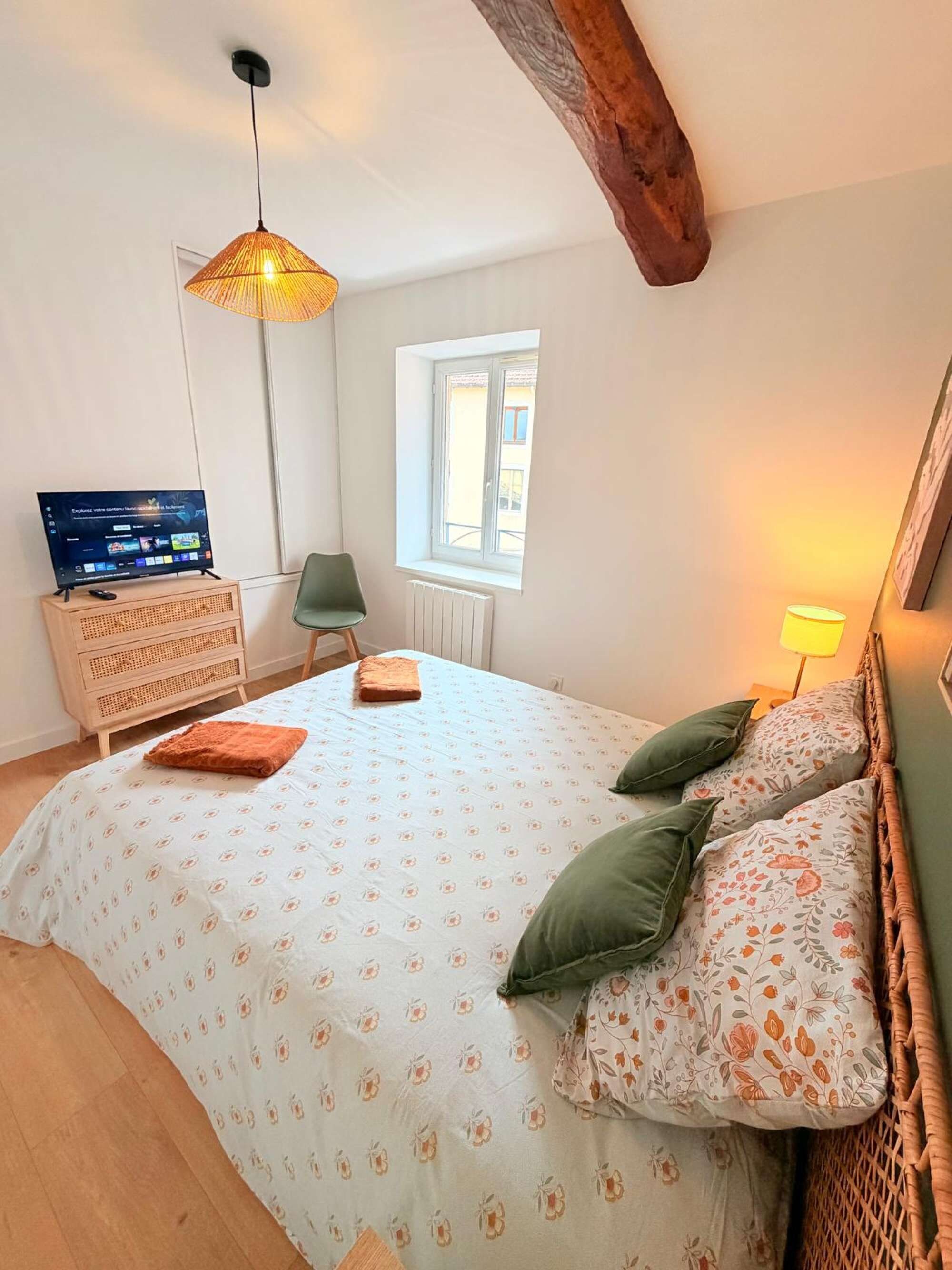 A bedroom with a bed, a television, a window, and a wooden ceiling beam.