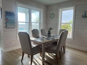 A dining area with a wooden table and chairs, a clock on the wall, and a view of the outdoors. - Mar+Sol by Liquid Life (Gulf Shores)