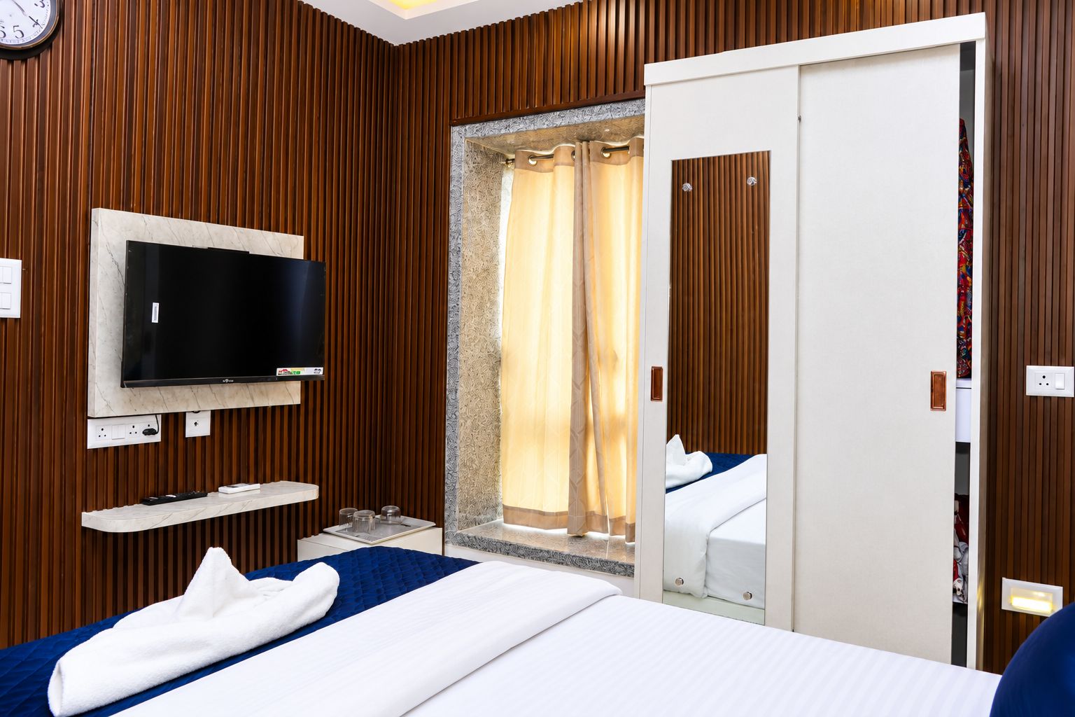 A hotel room with a bed, a TV mounted on the wall, a bathroom with a shower curtain, and a wooden wall paneling.