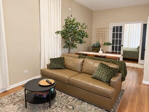 A living room with a brown leather sofa, a small round coffee table, a patterned rug, a potted plant, and a dining area with a table and chairs. - Chic Chicago Retreat, Stylish & Cozy Escape: 3 bedroom (Chicago)