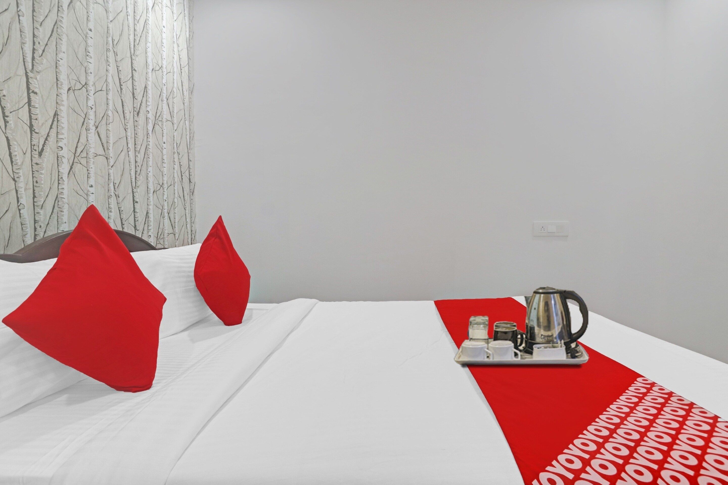 A hotel room with a bed, red pillows, a tray with a kettle, and a red and white patterned runner.