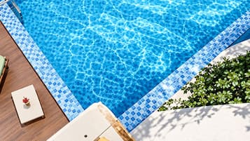 Una piscina rectangular con borde de mosaicos azules, rodeada por una terraza de madera y una zona de estar blanca.