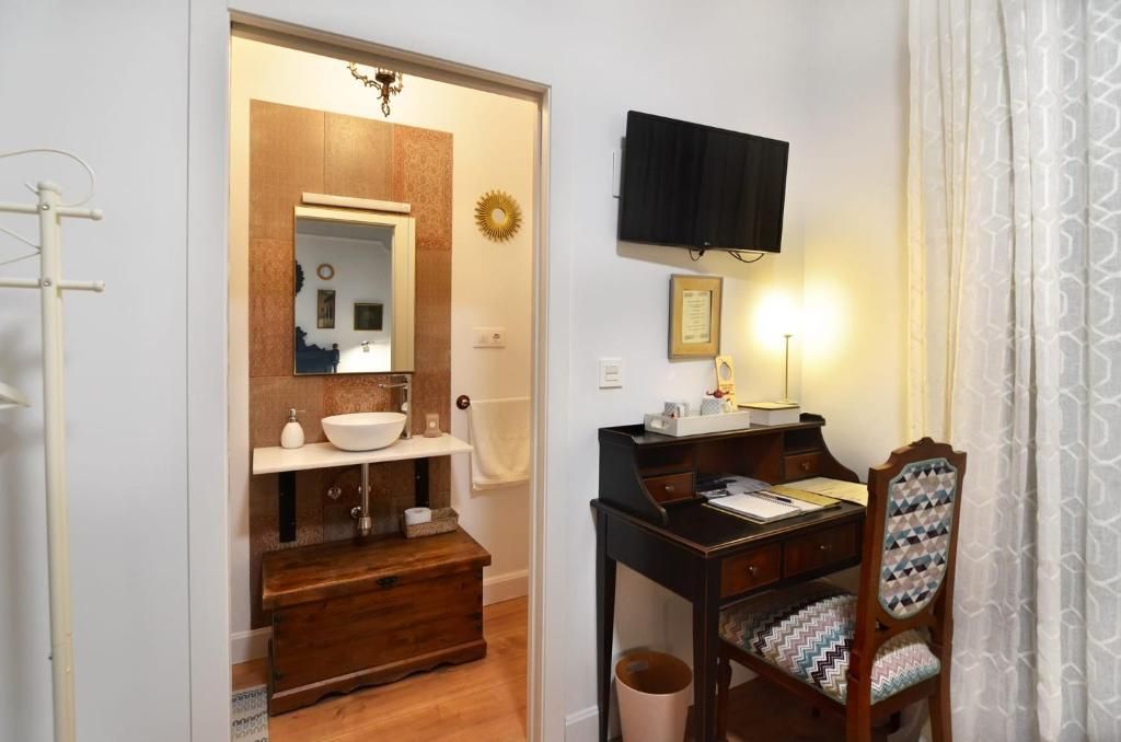 Un baño con tocador de madera, lavamanos, espejo, perchero para toallas, televisor montado en la pared y una silla con cojín estampado.