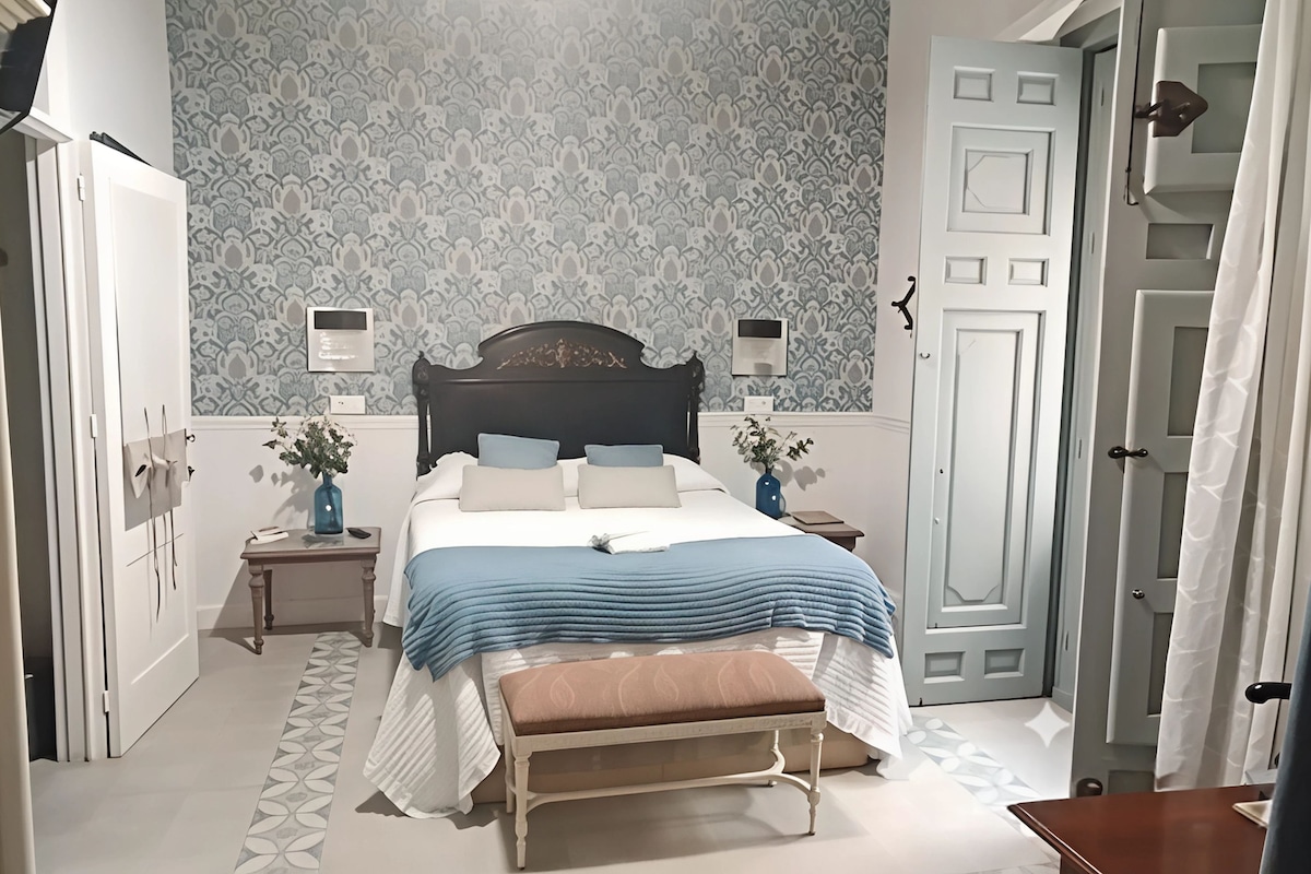Een slaapkamer met een bed, nachtkastjes, een bankje, een wandlamp en een deur.