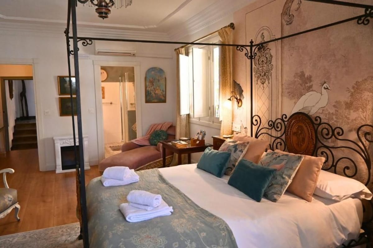 Een slaapkamer met een hemelbed, een zithoek met een fauteuil, een trap en een wanddecoratie van een pauw.