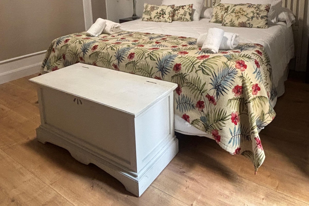 Een slaapkamer met een bed, een nachtkastje, een plant en een schilderij aan de muur.