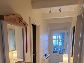A hallway with a door leading to a room, a window with curtains, and a mirror with a lamp. - Deichhof Apt. 21 (Dunsum)