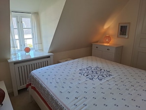 A bedroom with a bed, a bedside table, a lamp, a window with curtains, and a radiator. - Deichhof Apt. 21 (Dunsum)
