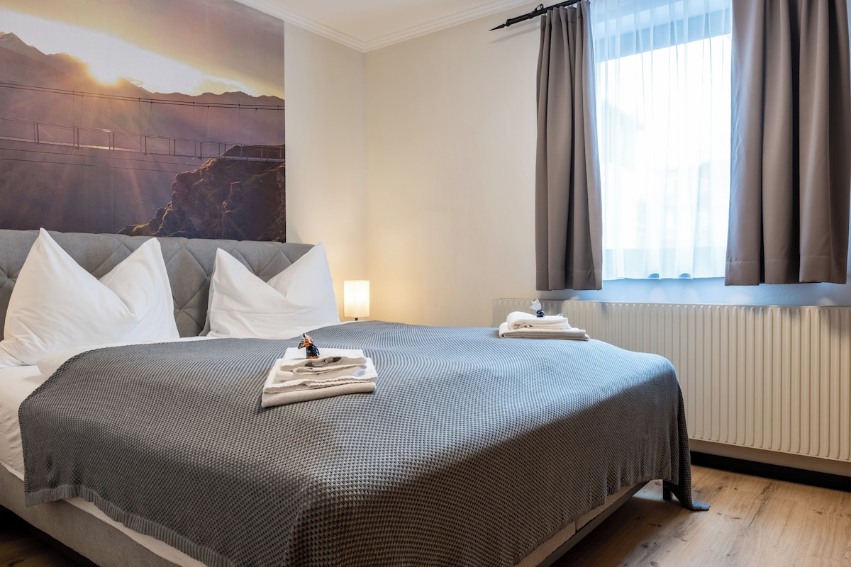 Ein Hotelzimmer mit Bett, Kissen, Nachttisch, Lampe, einem Fenster mit Vorhängen und einem Wandbild.