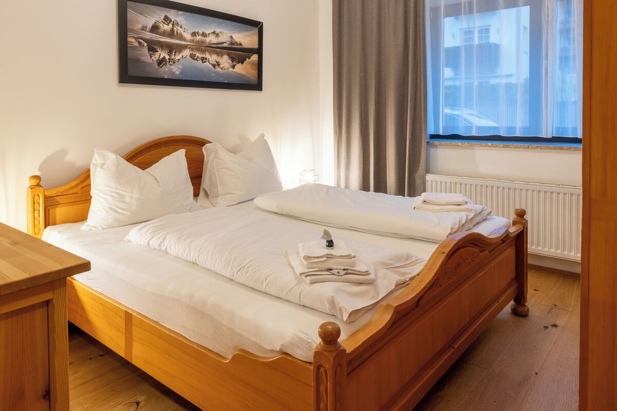 Ein ordentlich bezogenes Bett mit weißen Leinen und Kissen, ein hölzernes Kopfteil, ein Nachttisch, ein Fenster mit Vorhängen und ein gerahmtes Bild an der Wand.