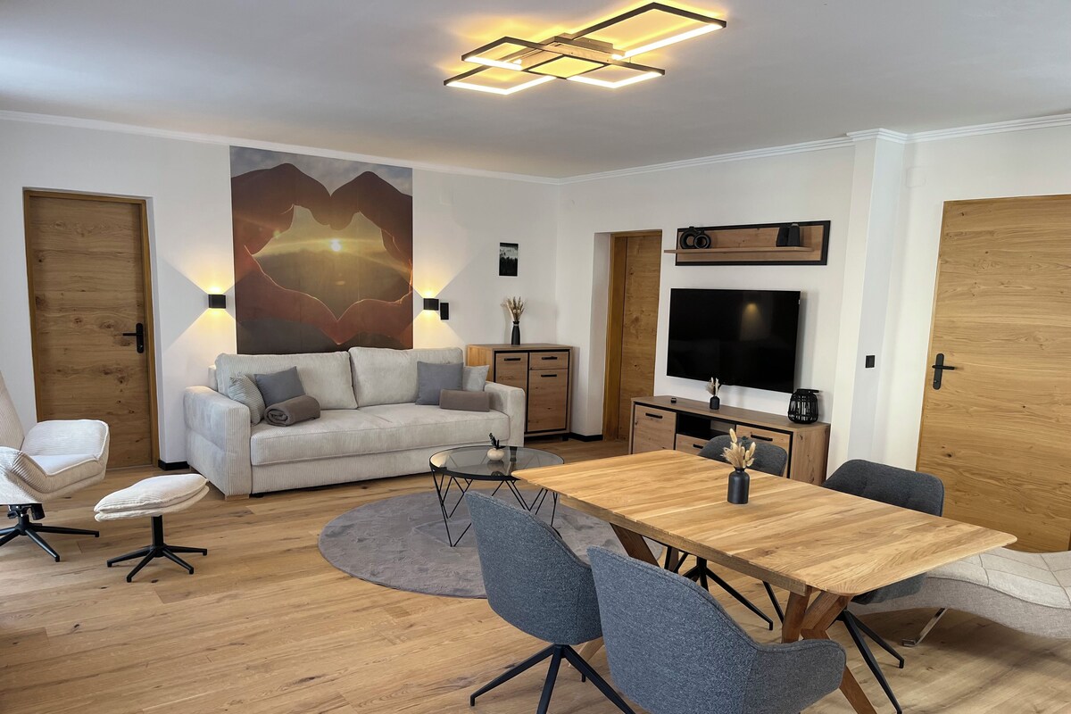 Ein modernes Wohnzimmer mit einer Couch, einem Esstisch und Stühlen. Ein Fernseher ist an der Wand montiert und es gibt ein Holzschränkchen.