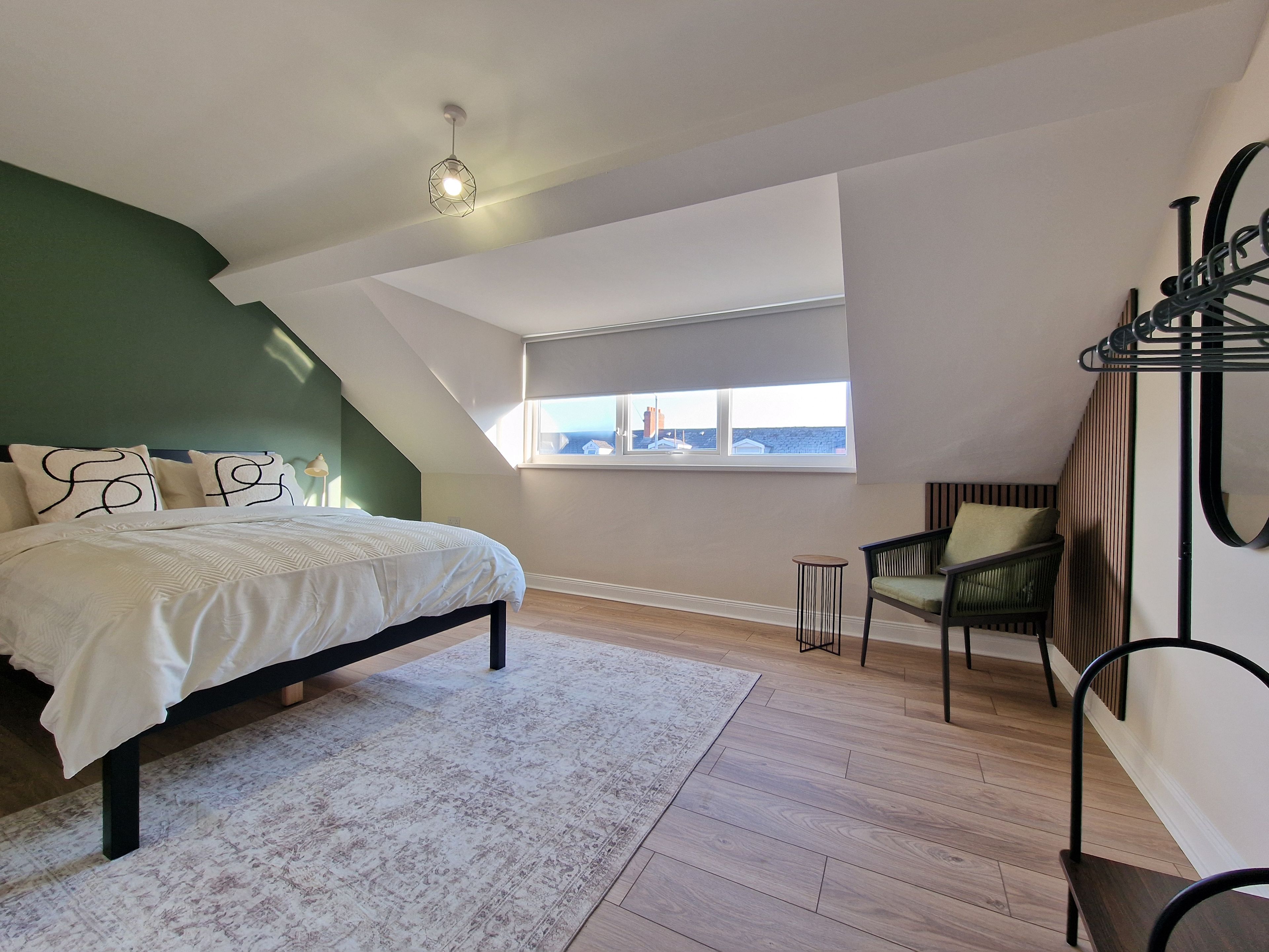 A bedroom with a sloped ceiling, a bed with a white bedspread, a small round table, a chair, and a view of the outside through a window.