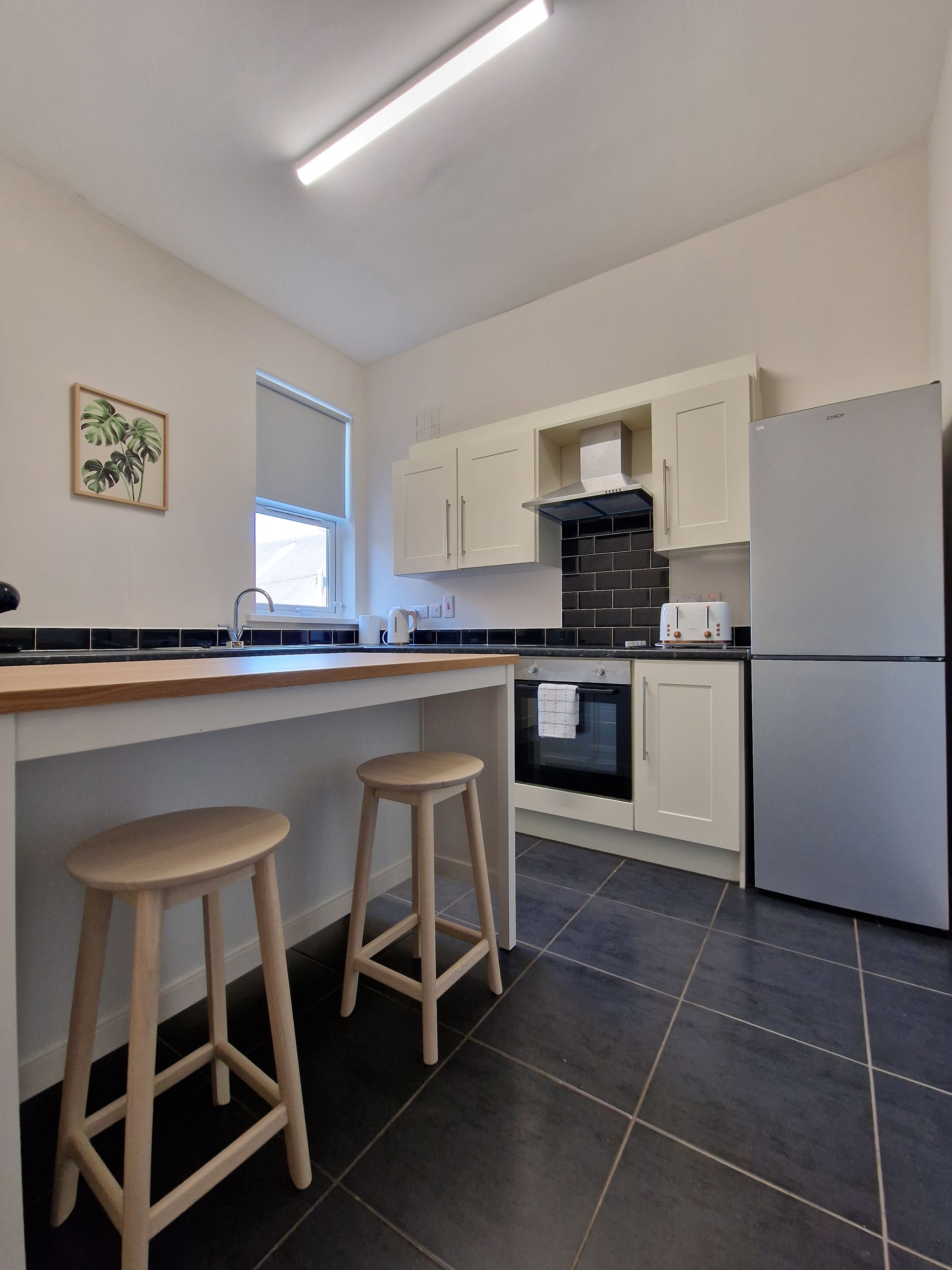 A modern kitchen with a central island, two bar stools, a refrigerator, and a window with blinds.