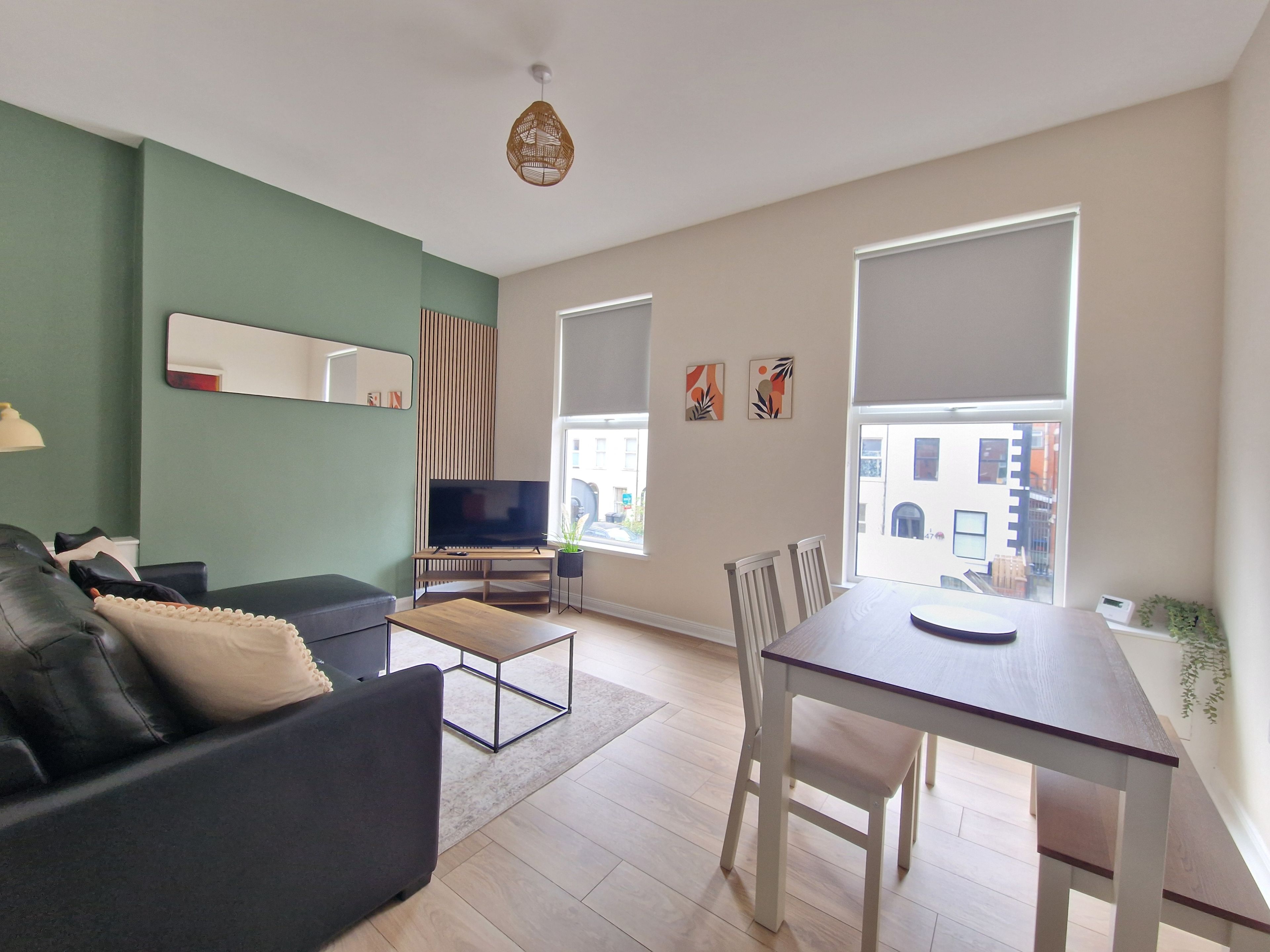 A modern living room with a sofa, a TV on a stand, a dining table with chairs, and large windows with blinds.