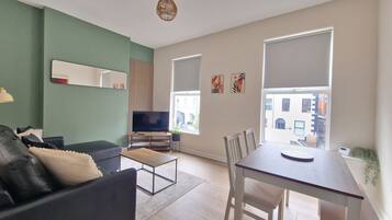 A modern living room with a sofa, a TV on a stand, a dining table with chairs, and large windows with blinds.