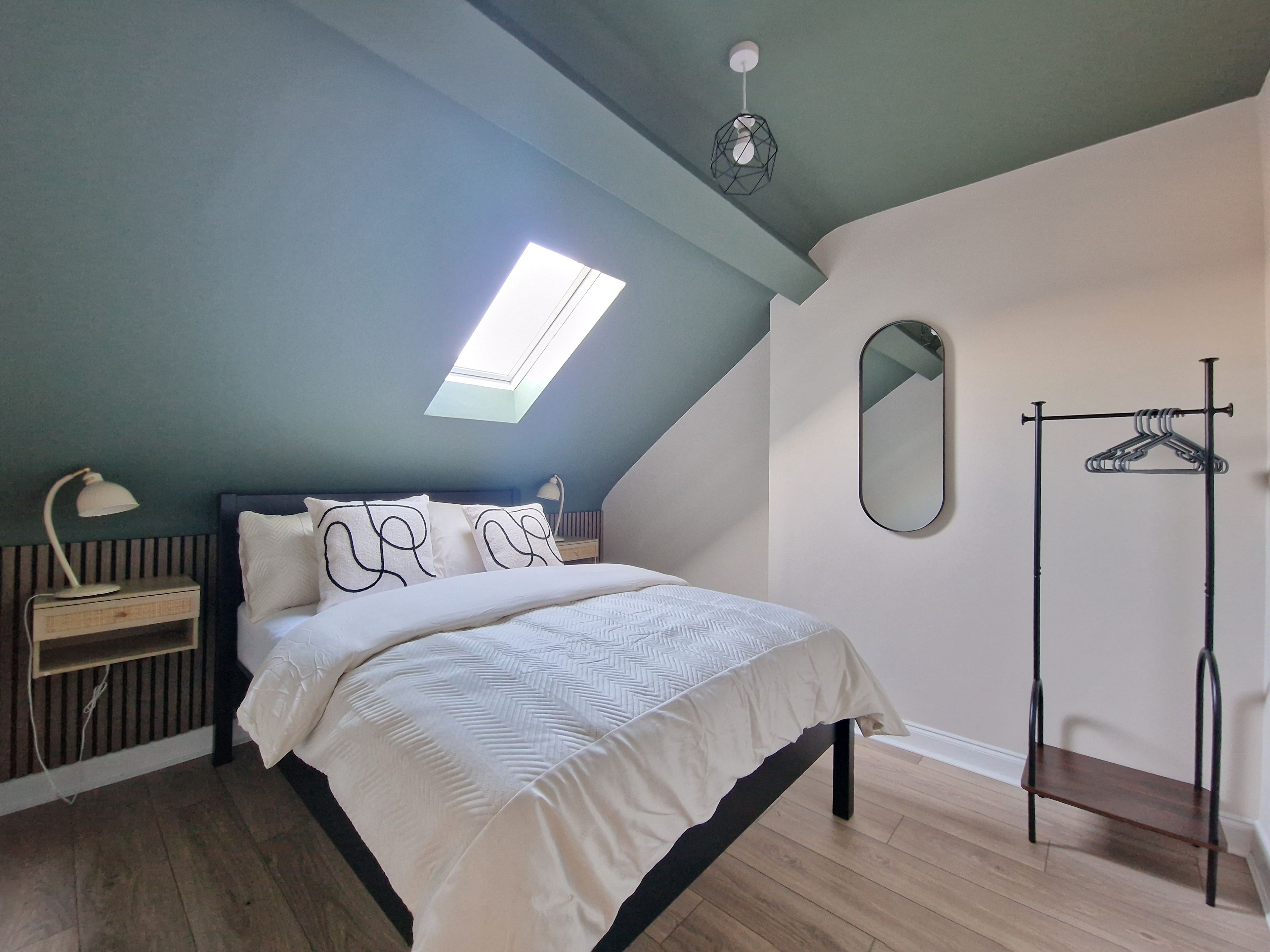 A bedroom with a sloped ceiling, a skylight, a bed with white bedding, a bedside table with a lamp, a mirror, and a clothing rack.