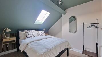 A bedroom with a sloped ceiling, a skylight, a bed with white bedding, a bedside table with a lamp, a mirror, and a clothing rack.