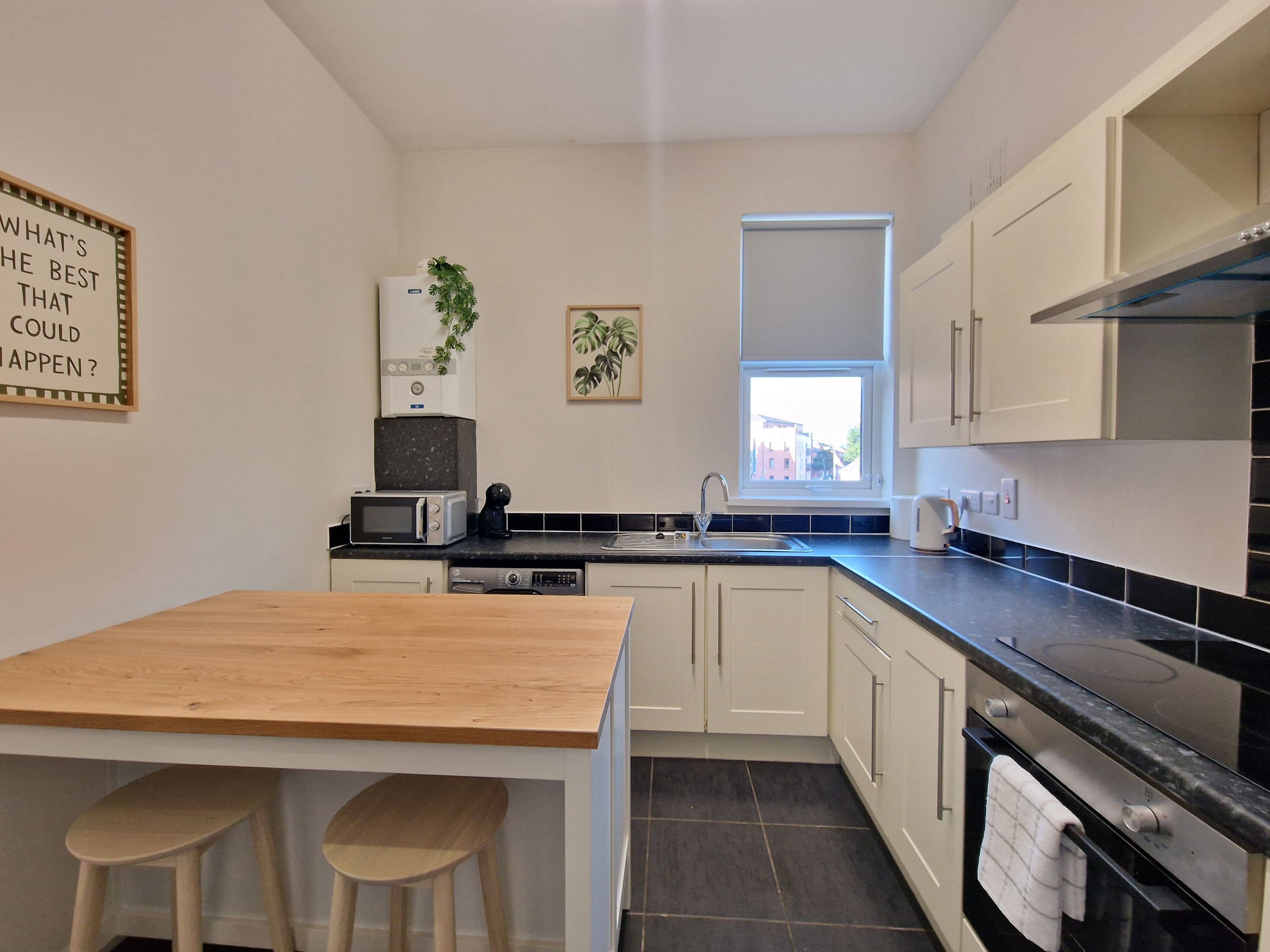 A compact kitchen with a wooden table, two stools, a microwave, a coffee maker, and a window with a view.