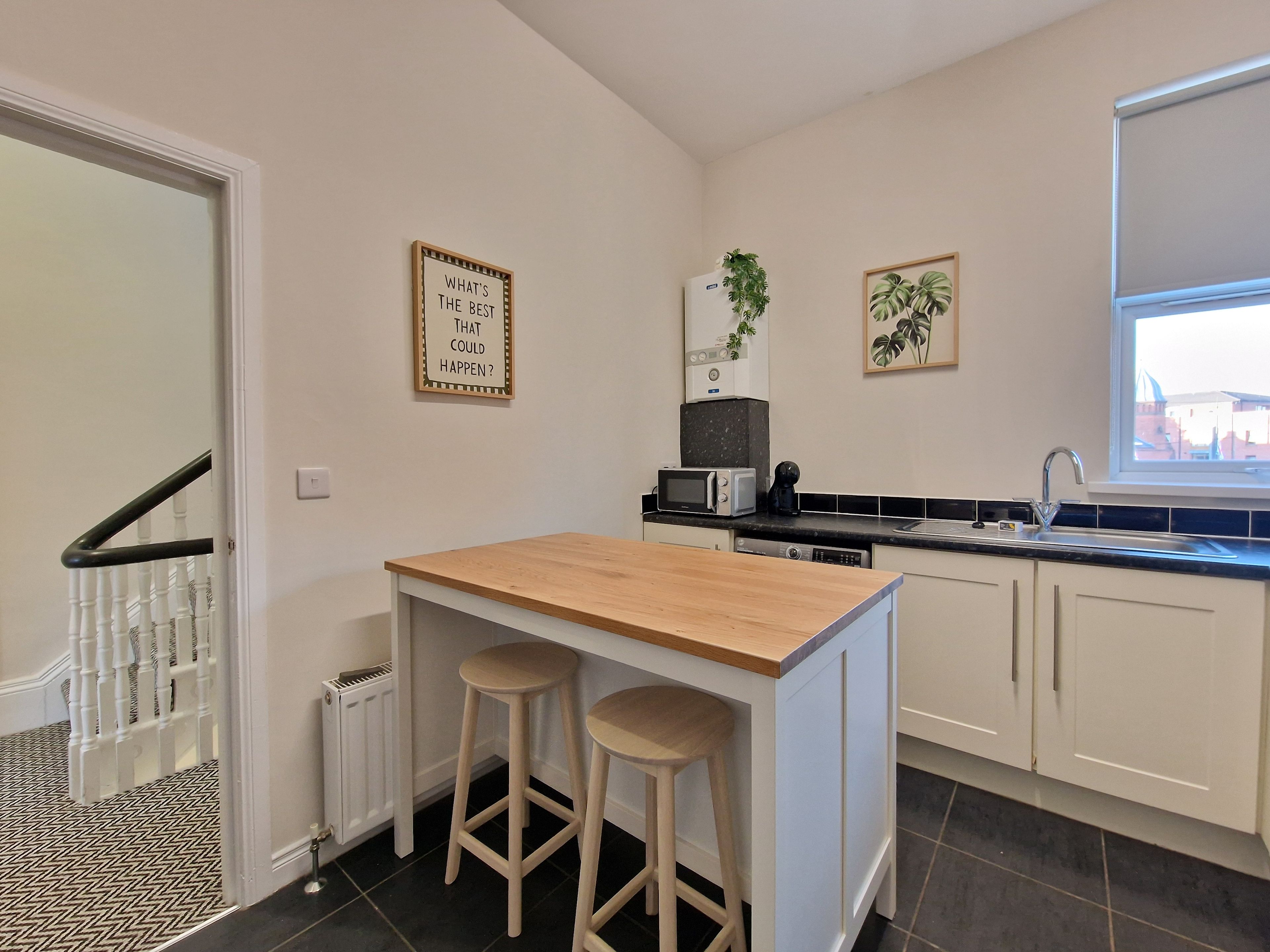A compact kitchen with a wooden island, two stools, a microwave, a coffee maker, and a sink.