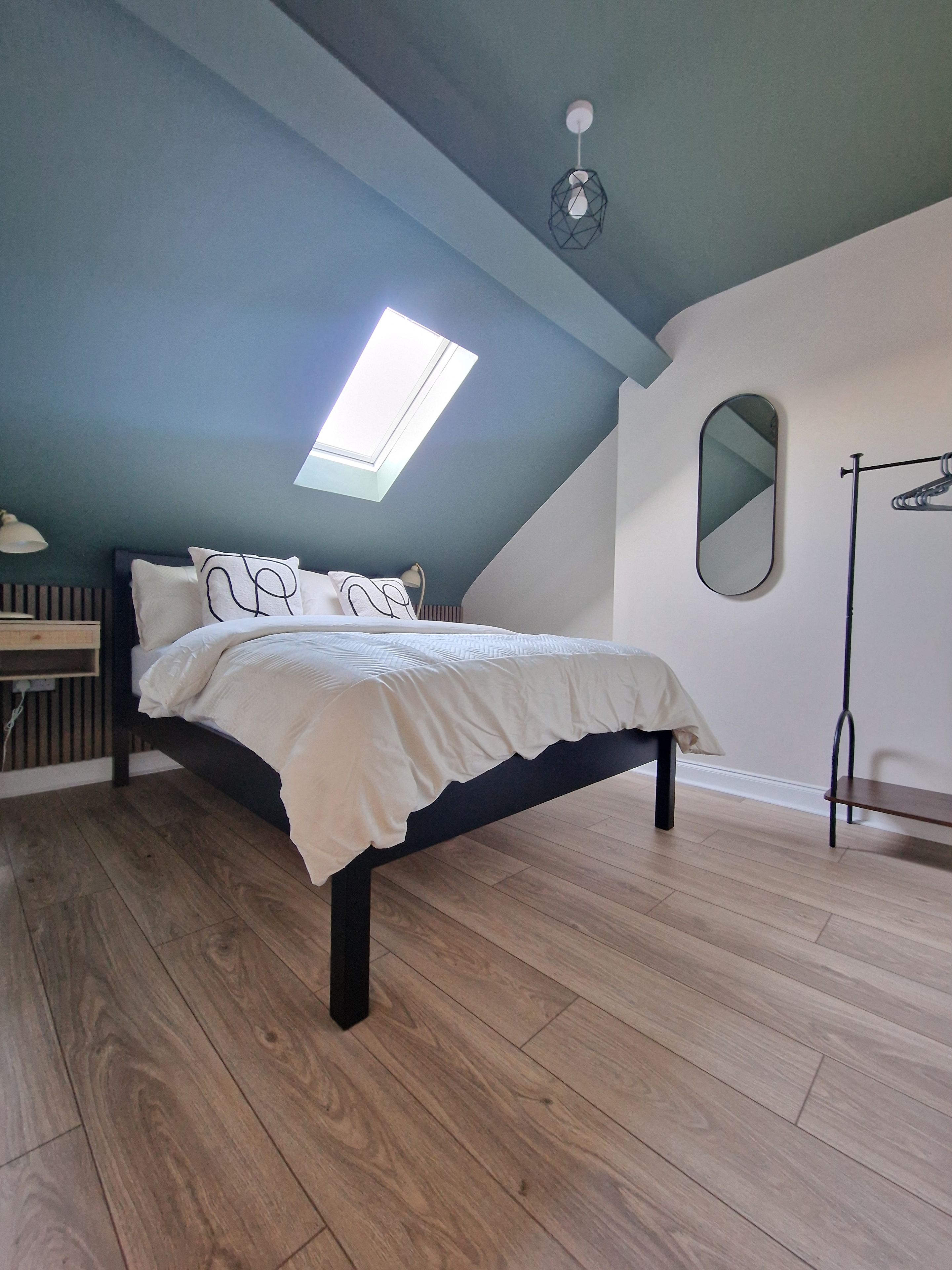 A bedroom with a sloped ceiling, a skylight, a bed with white bedding, a wooden floor, a bedside table, and a hanging light fixture.