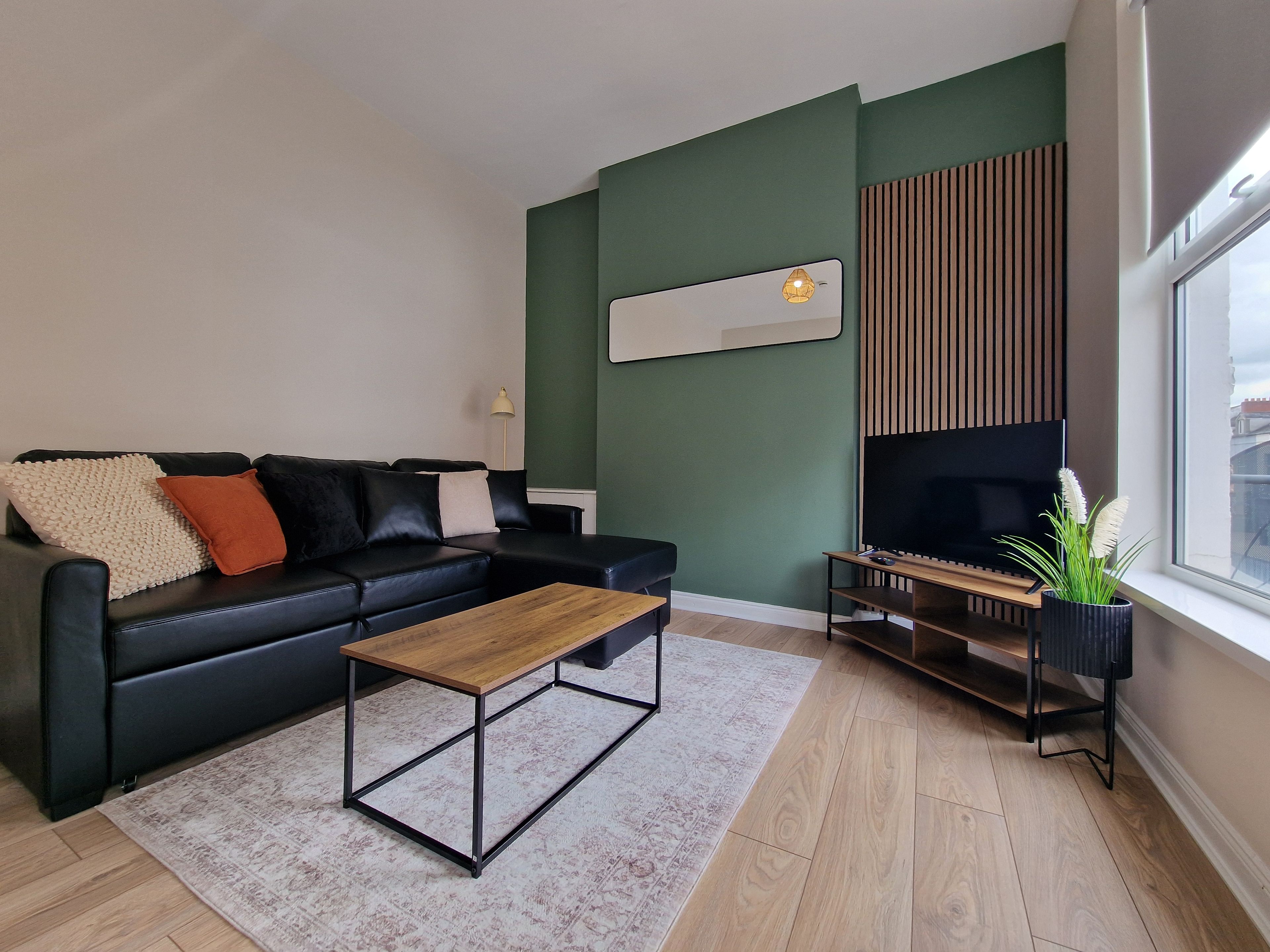 A modern living room with a black leather sofa, a wooden coffee table, a flat-screen TV on a wooden stand, and a large window.