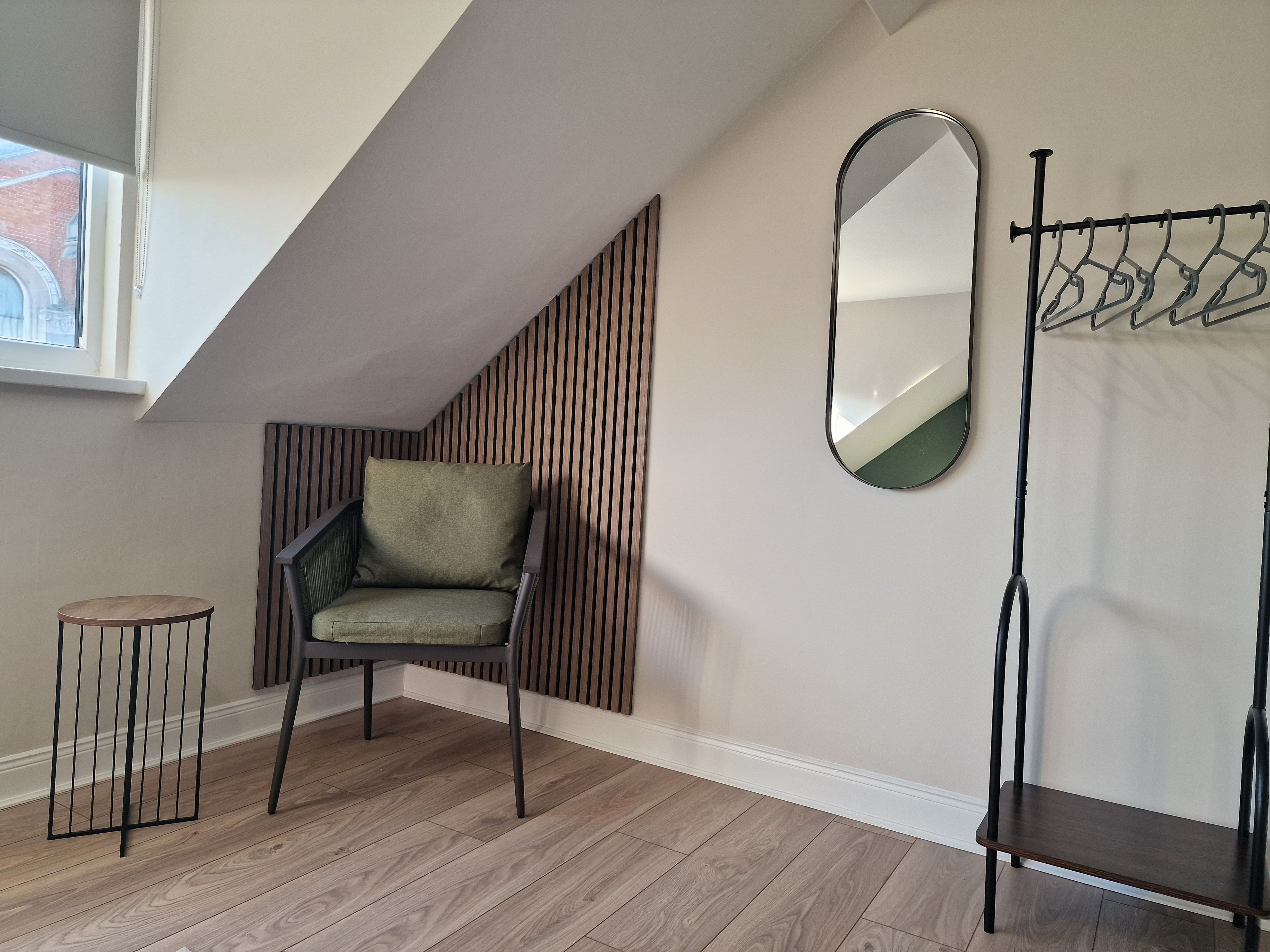 A room with a sloped ceiling, a green upholstered chair, a small round table, a mirror, and a coat rack.