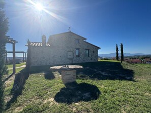 Una casa in pietra con tetto spiovente, un piccolo giardino e una vista aperta sul paesaggio circostante.