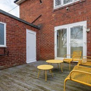 Un bâtiment en briques avec une porte et des fenêtres blanches, un patio couvert avec des chaises jaunes et une table.