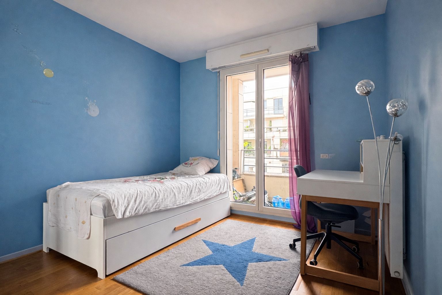 Une chambre avec un lit simple, un bureau, une chaise et un tapis orné de motifs en forme d’étoiles.
