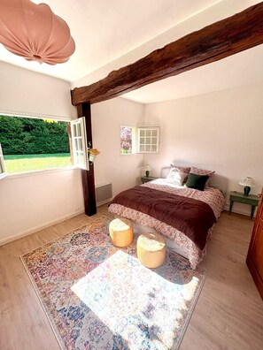 A bedroom with a large bed, a window with a view of greenery, a wooden ceiling beam, and a patterned rug. - Normandy farmhouse with a fireplace and a 2,800 m² enclosed garden. Pets welcome (Heudicourt)