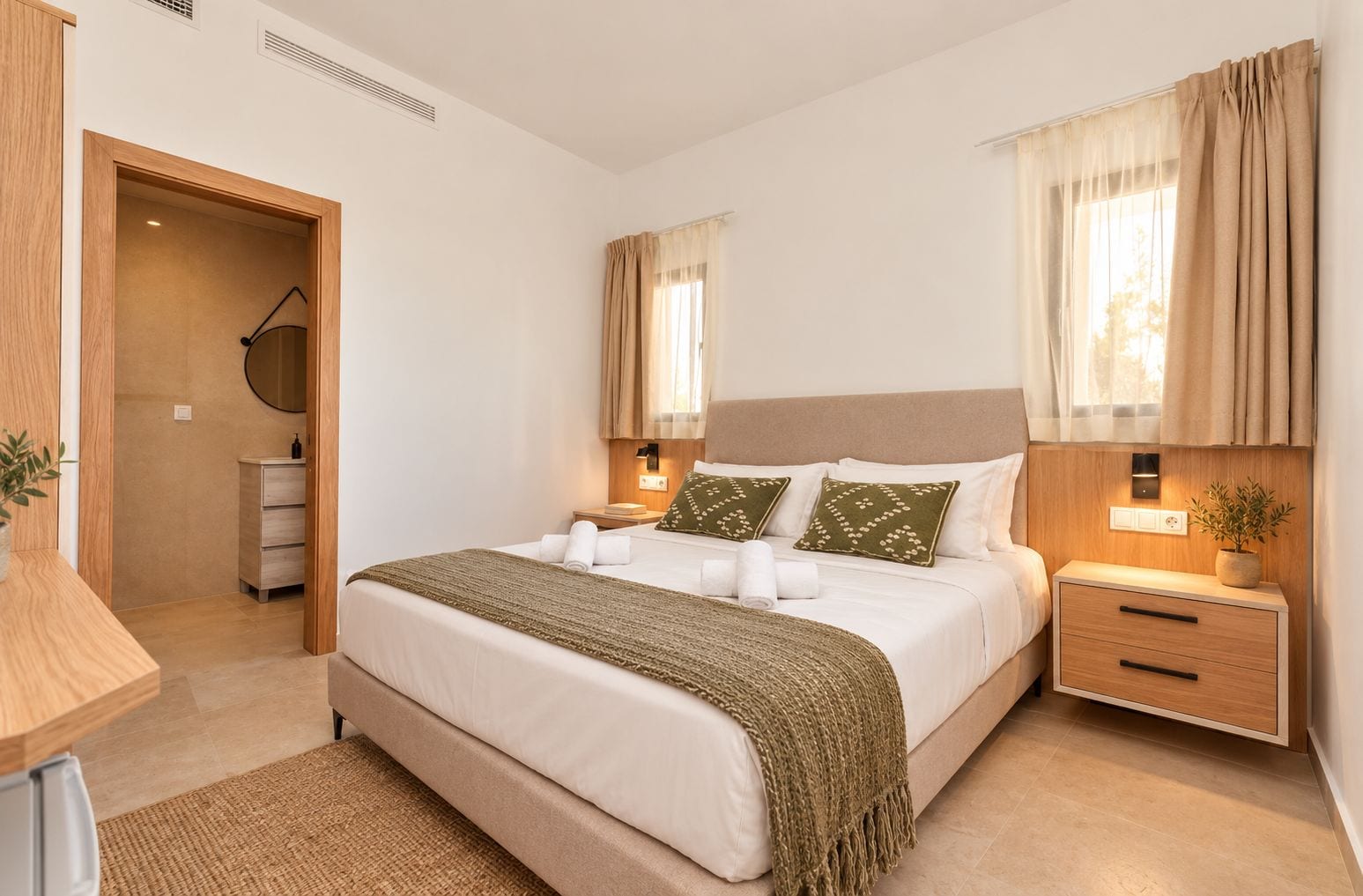 Quarto de hotel com uma cama grande, mesas de cabeceira, um guarda-roupa de madeira e uma pequena escrivaninha com uma planta.