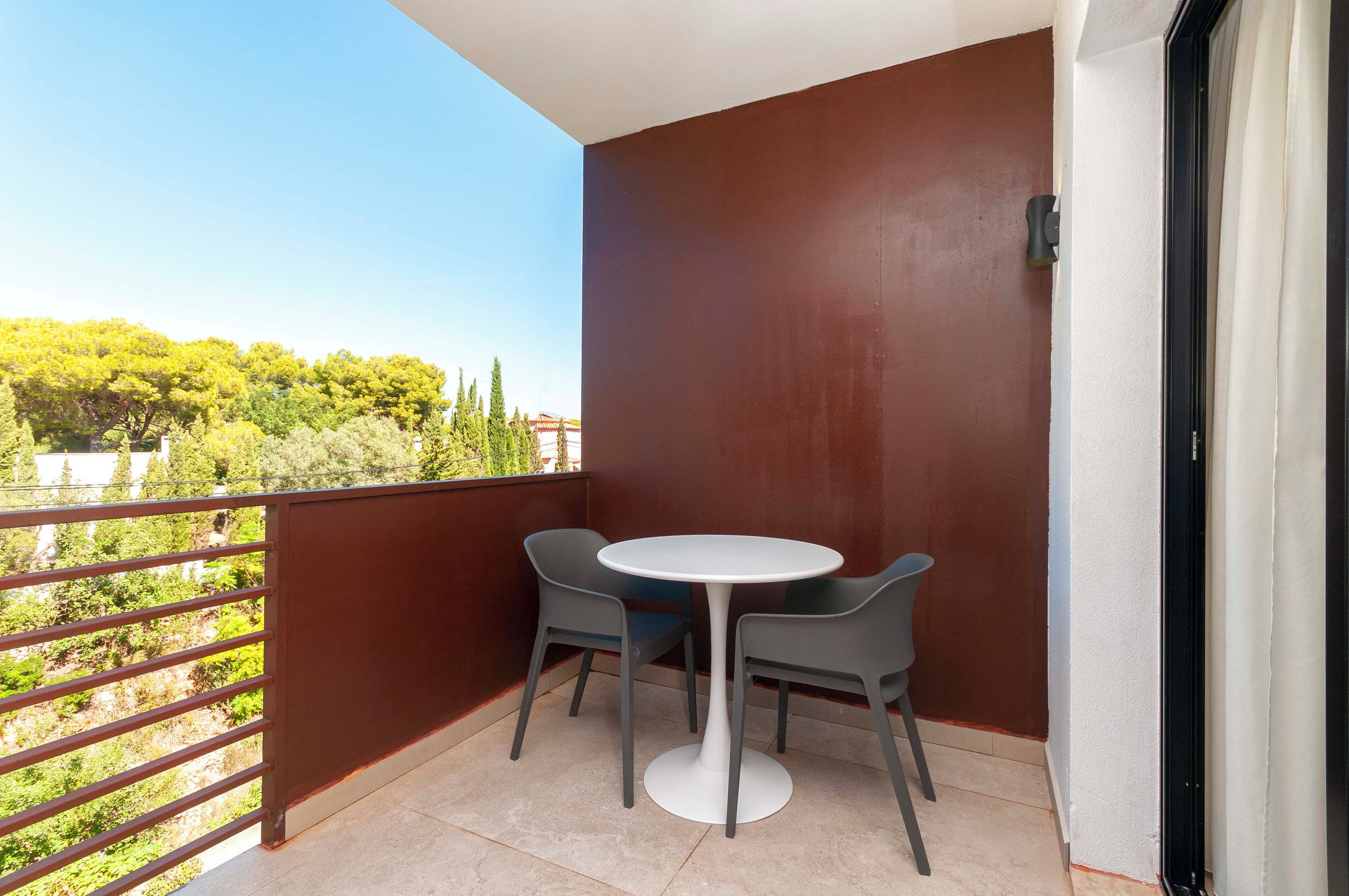 Un balcón con una mesa blanca y dos sillas, una pared roja y vista a zonas verdes.