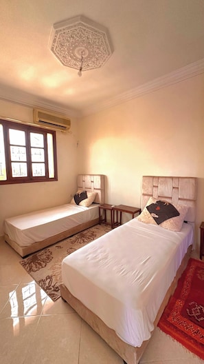 Quarto de hotel com duas camas, ventoinha no teto, aparelho de ar condicionado e janela com vidro decorativo.