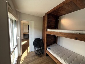 Uma cama beliche com colchão e cabeceira de madeira em quarto com piso de madeira e porta que dá para outro cômodo.