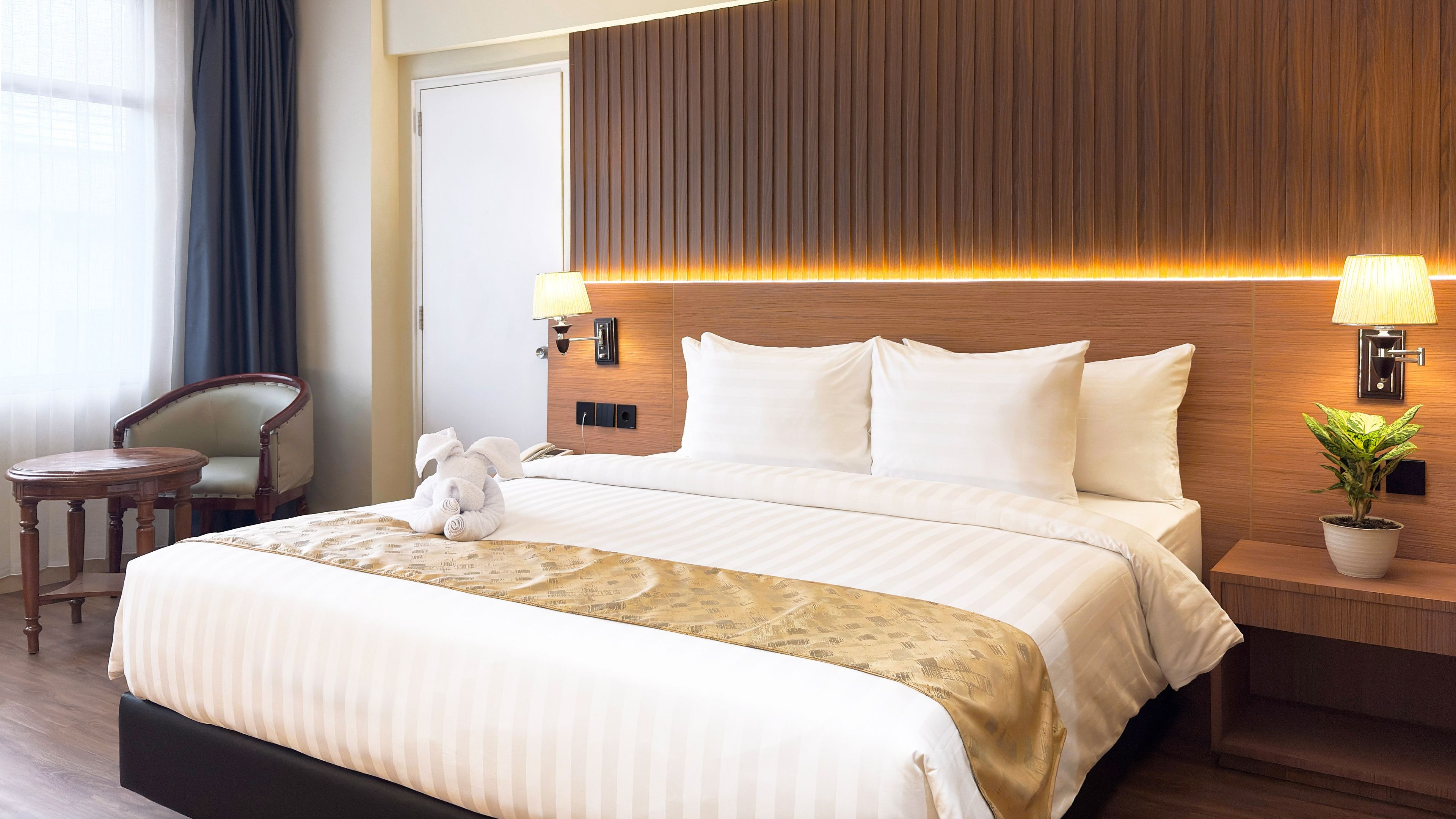 A hotel room with a large bed, two bedside lamps, a wooden headboard, a small round table, a chair, and a potted plant.