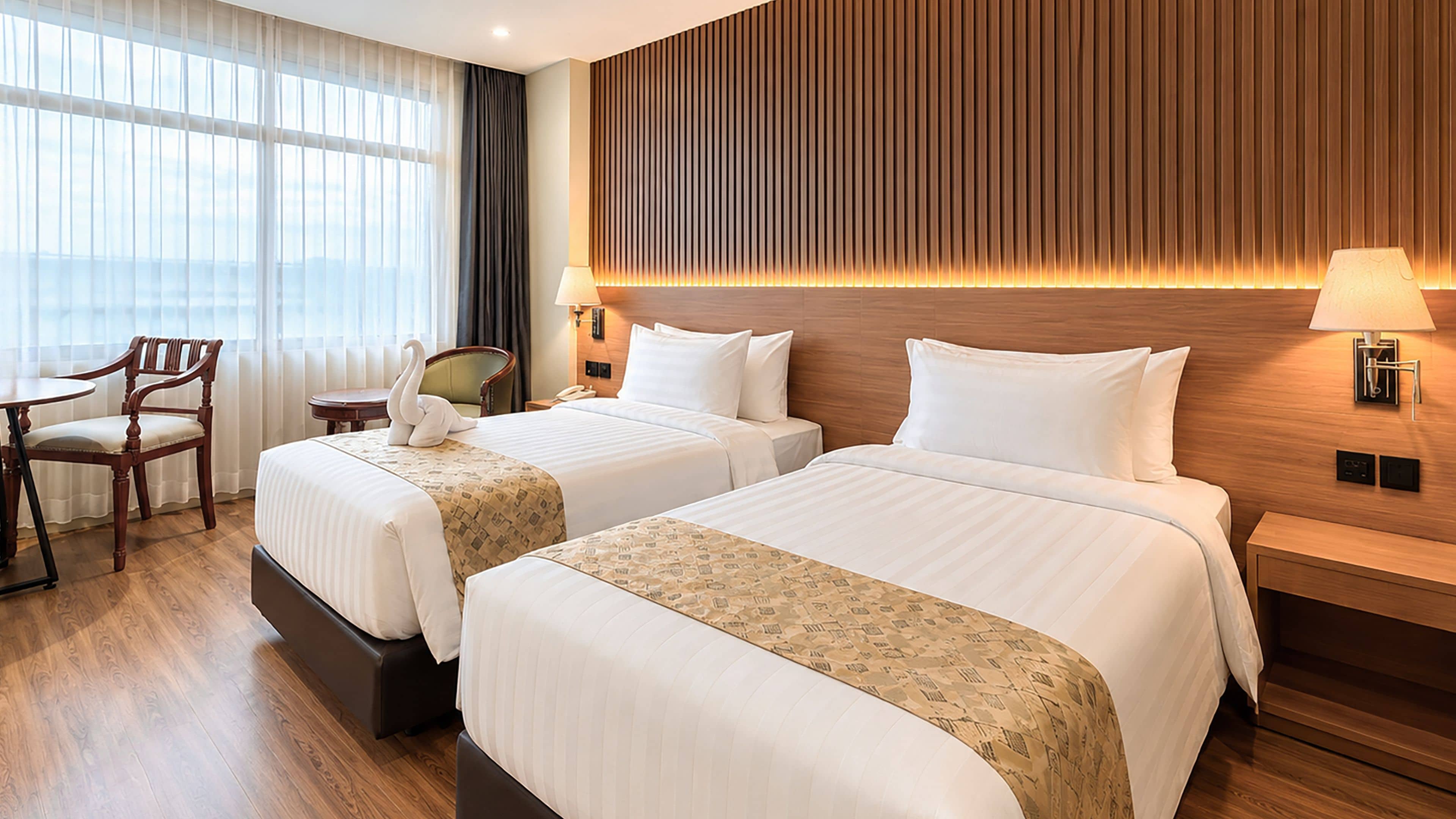 A hotel room with two beds, a sitting area with a chair, a wooden headboard, and a view of the city.
