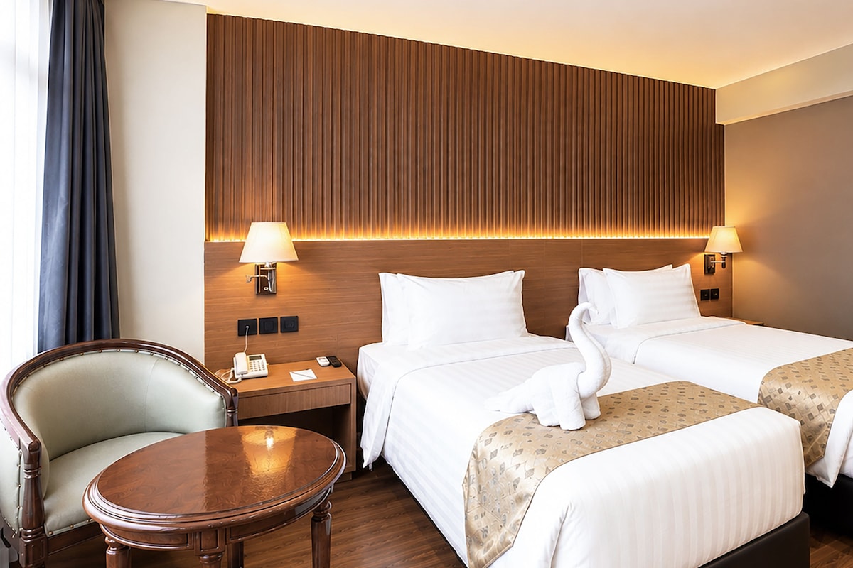 Een hotelkamer met twee bedden, een houten hoofdeinde, nachtlampjes, een klein rond tafeltje en een stoel.