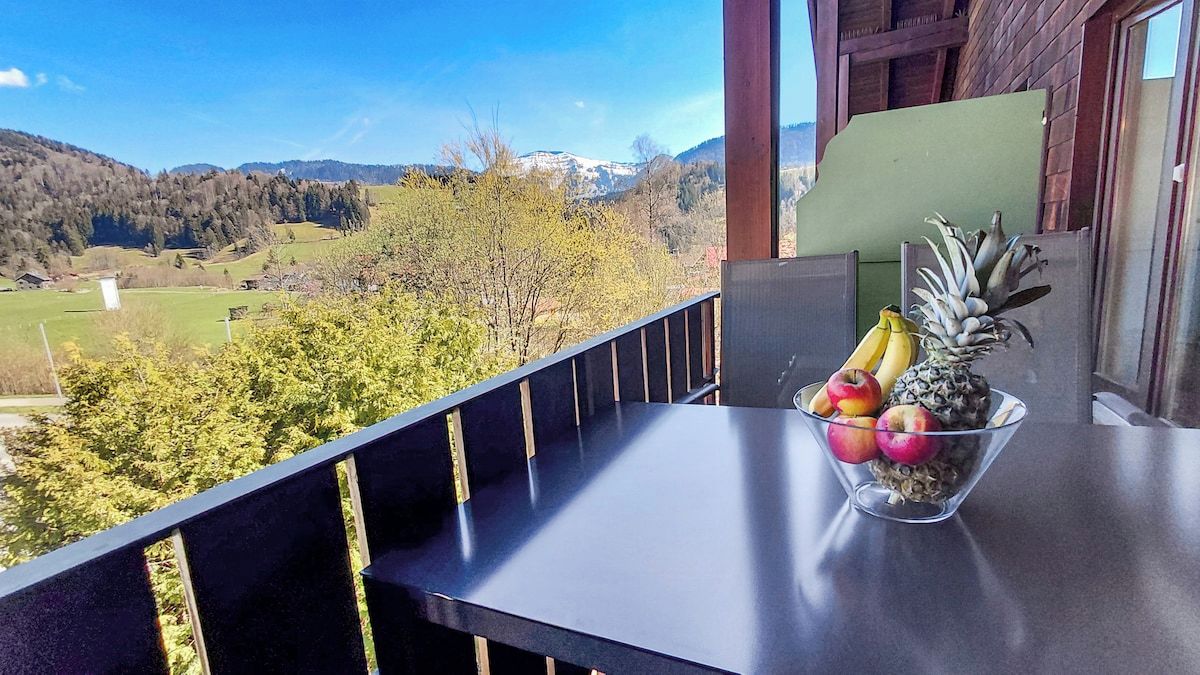 Een balkon met vrij uitzicht op een bergachtig landschap, een schaal fruit op de tafel en een houten leuning.