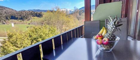 Een balkon met vrij uitzicht op een bergachtig landschap, een schaal fruit op de tafel en een houten leuning.