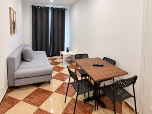 A modern living room with a grey sofa, a wooden dining table with four chairs, a small white table with a plant, and a window with black curtains. - Settle Into Vienna 2BR near Westbahnhof (Vienna)