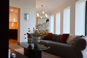 Un salon moderne avec un canapé gris, une table basse noire et un vase contenant des fleurs séchées. Un tableau est accroché au mur et un luminaire est fixé au plafond.