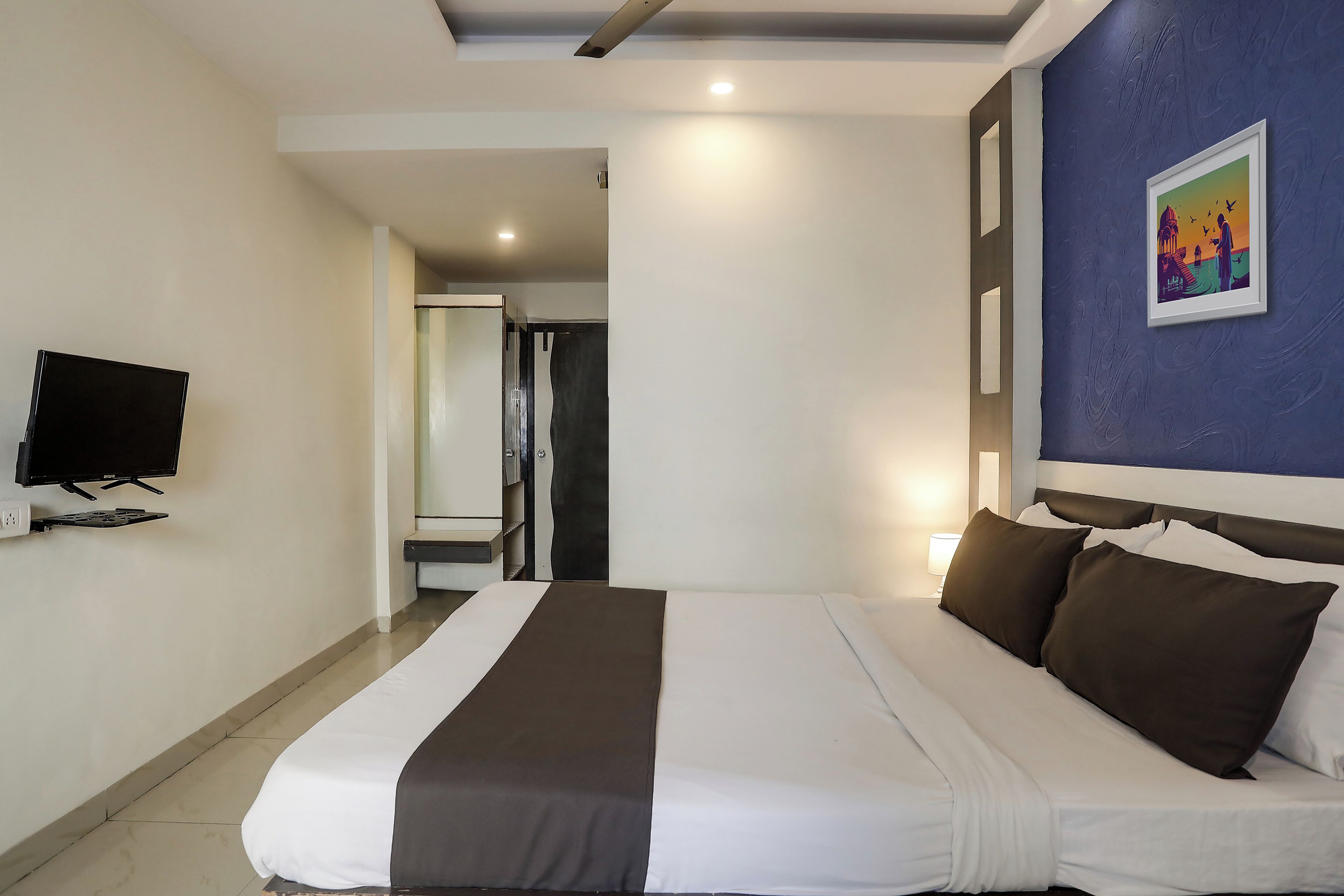 A hotel room with a bed, a television, a painting on the wall, and a door leading to another room.