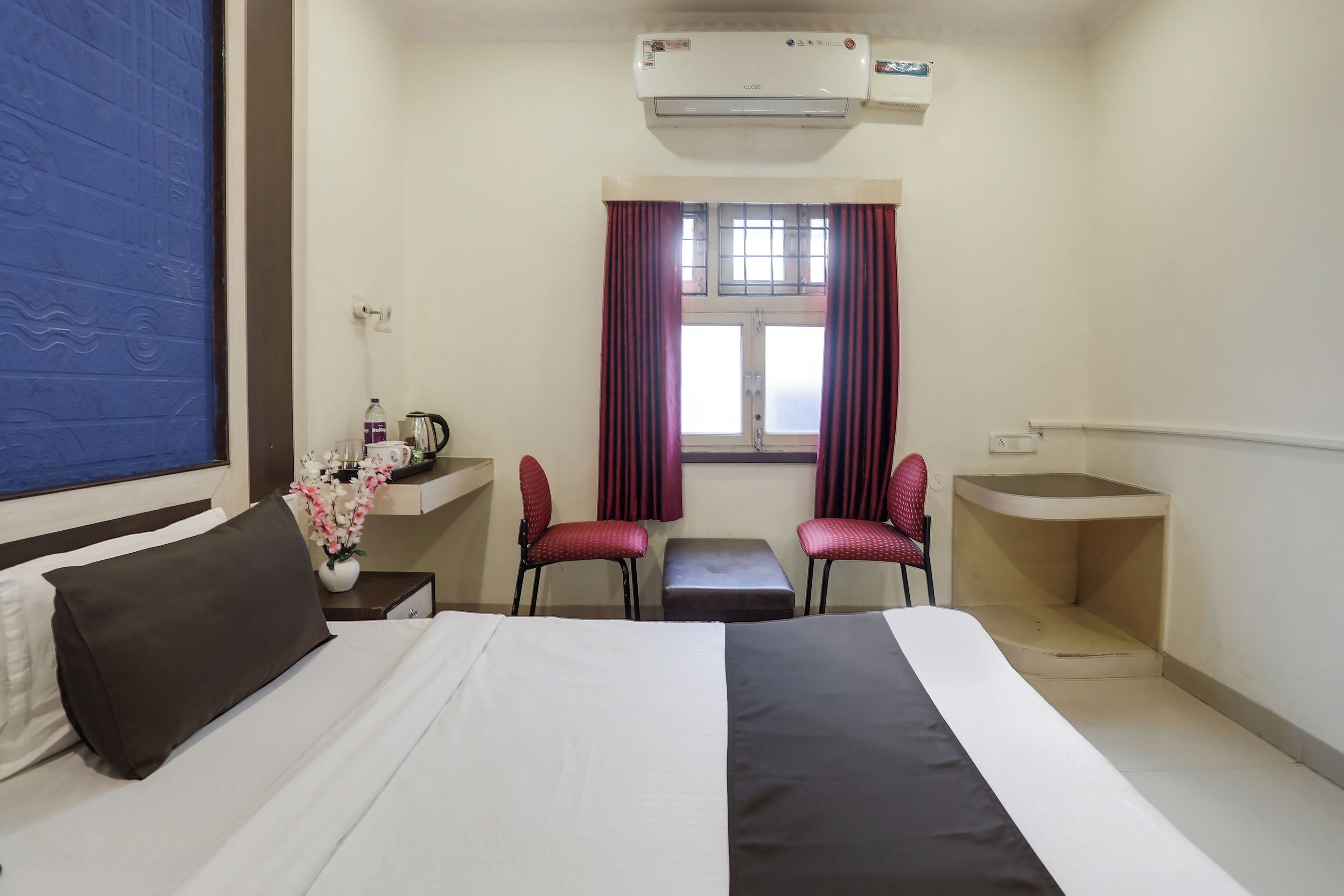 A hotel room with a bed, a desk, two chairs, a window with curtains, and an air conditioning unit.